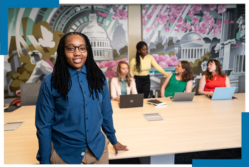GSEHD student smiles at camera, other students work on laptops at table in background