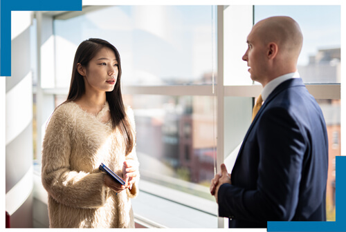 two GSEHD students talk with each other in front of large windows with campus in the background